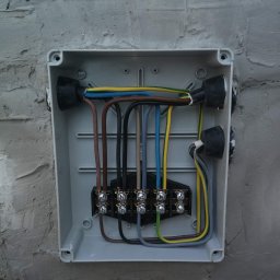 Elektro-Lasiu - Szara skrzynka elektryczna z podłączonymi przewodami o różnych kolorach zamontowana na szorstkiej, otynkowanej ścianie. Widoczne czarne gumowe uszczelki wokół otworów na przewody.