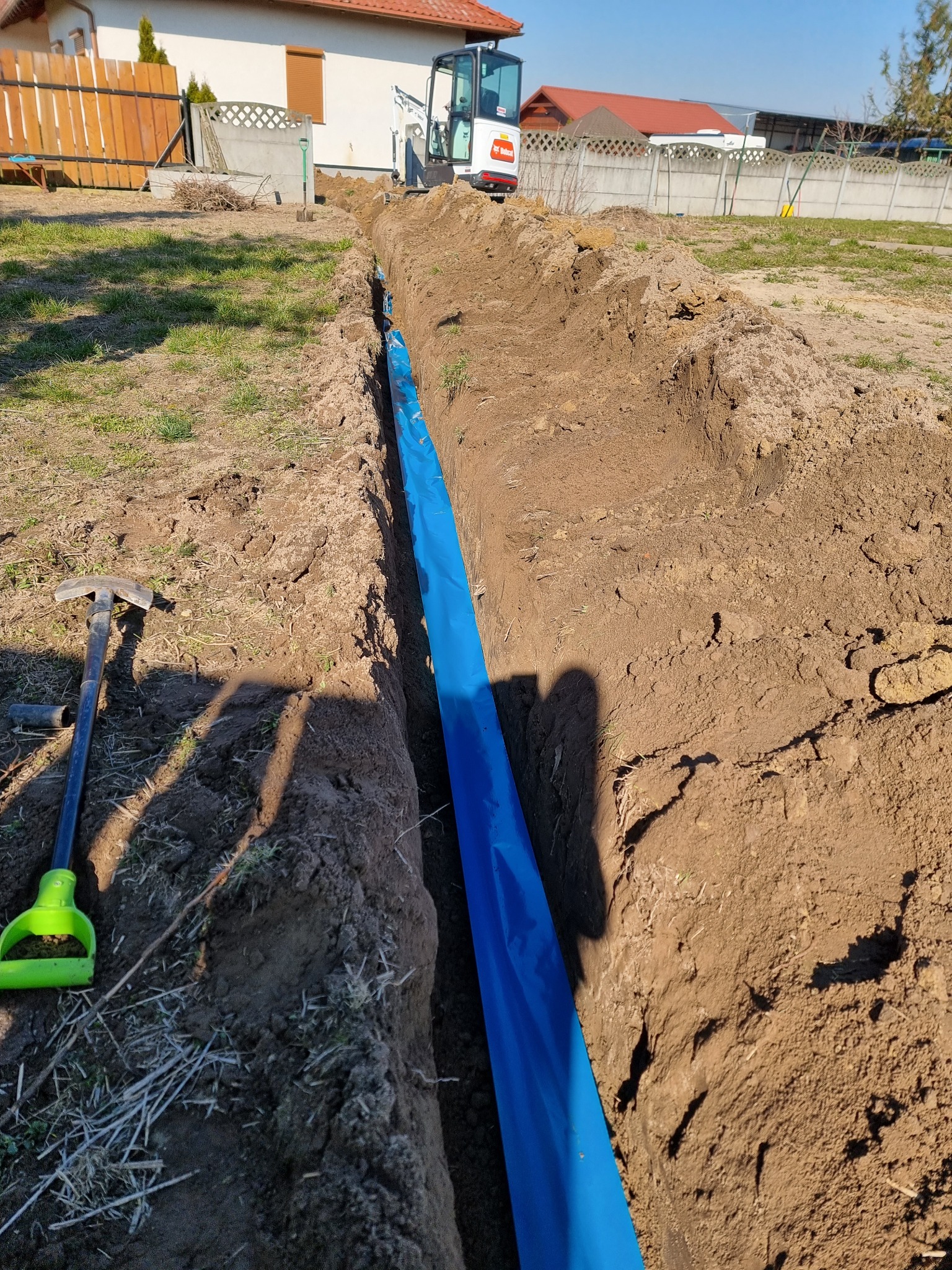 Wykop w ziemi wyłożony niebieską folią ochronną, w tle minikoparka Bobcat i zabudowa jednorodzinna, obok wykopu leży szpadel z zieloną rączką.