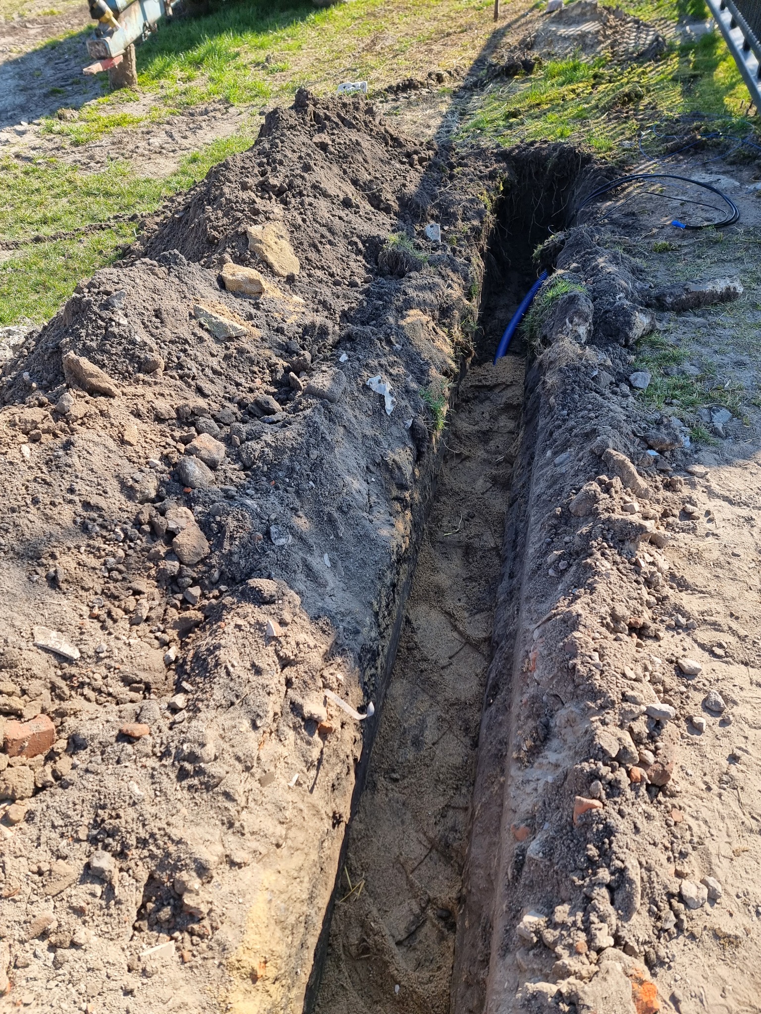 Wykop w ziemi z ułożonym kablem w niebieskiej rurze karbowanej, widoczne ułożone warstwy ziemi i gruzu po wykopie.