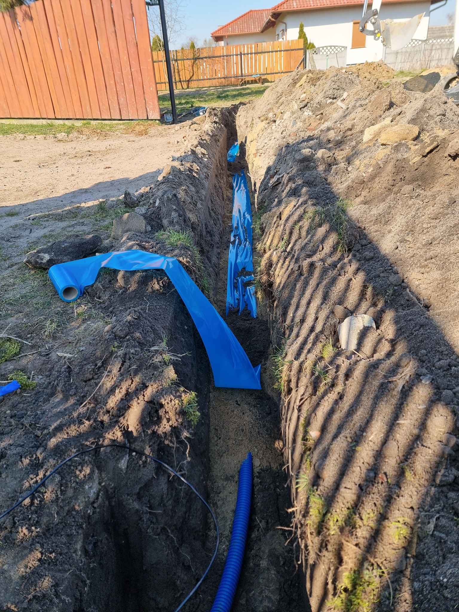 Wykop w ziemi z niebieską rurą karbowaną i folią ochronną, przygotowany pod ułożenie kabli elektrycznych na posesji jednorodzinnej w słoneczny dzień.