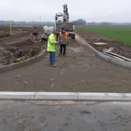 Dwóch robotników wyrównuje kruszywo na przygotowywanej drodze, obok ułożone krawężniki, w tle ciężarówka i koparka na placu budowy.