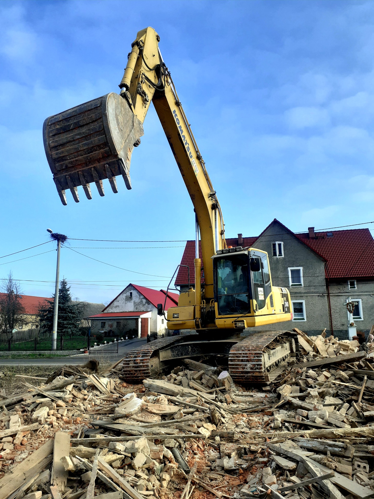Żółta koparka Komatsu na gruzowisku po rozbiórce budynku, w tle domy z czerwonymi dachami i słup z gniazdem bocianim na tle błękitnego nieba.