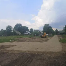 Przygotowany teren pod budowę z wyznaczonymi fundamentami, widoczny żółty traktor i pracownik z niwelatorem optycznym.