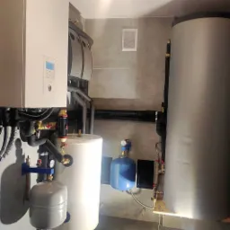 Złożony system instalacji grzewczej w pomieszczeniu z szarymi płytkami, widoczne zbiorniki, rury i zawory w różnych kolorach.