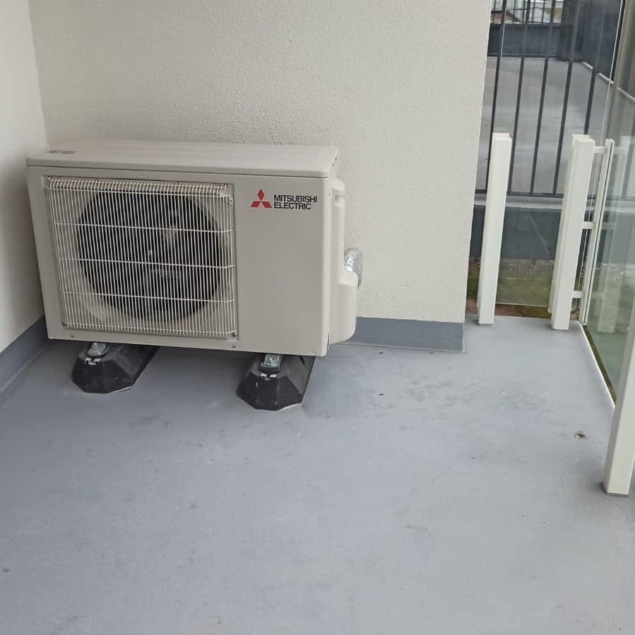 Zewnętrzna jednostka klimatyzacji Mitsubishi Electric zamontowana na czarnych podstawkach na szarym balkonie z metalową barierką w tle.