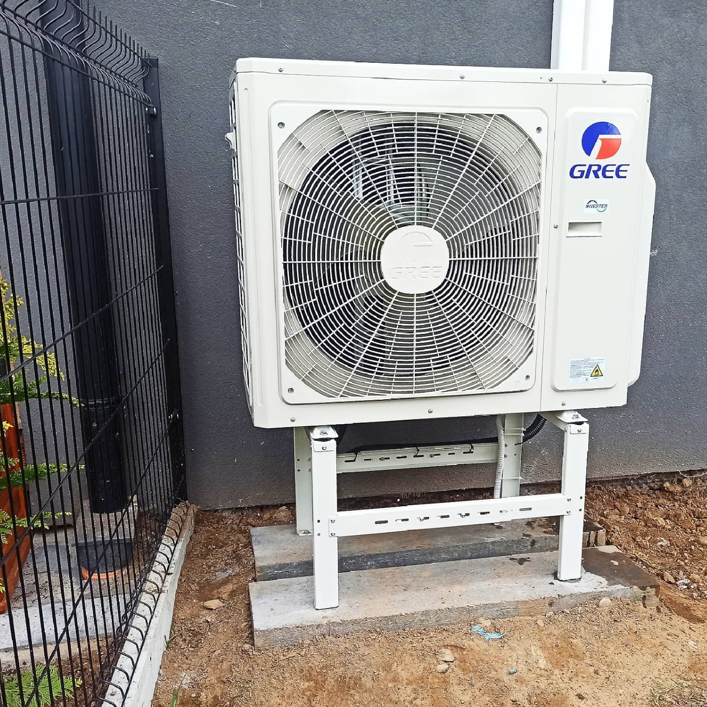 Zewnętrzna jednostka klimatyzacji GREE Inverter zamontowana na białym stelażu przy szarej ścianie budynku, widoczna czarna siatka ogrodzeniowa i ziemia.
