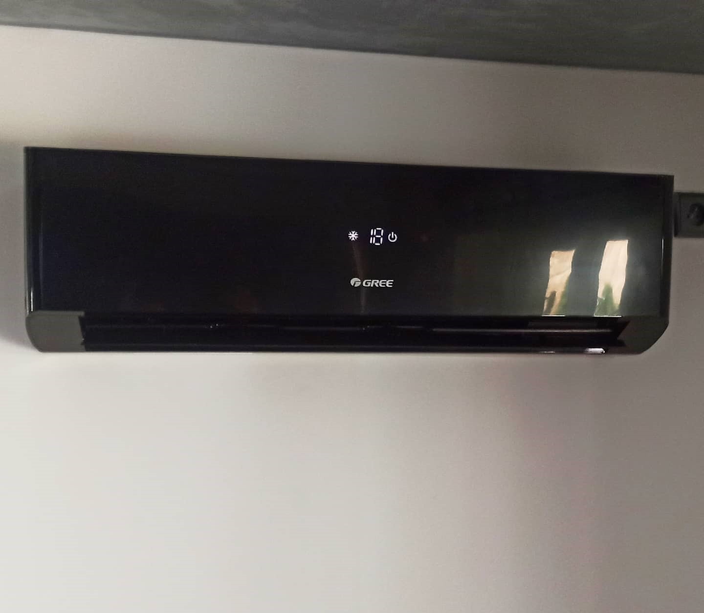 Czarny klimatyzator marki Gree zamontowany na białej ścianie, wyświetlający temperaturę 18 stopni.