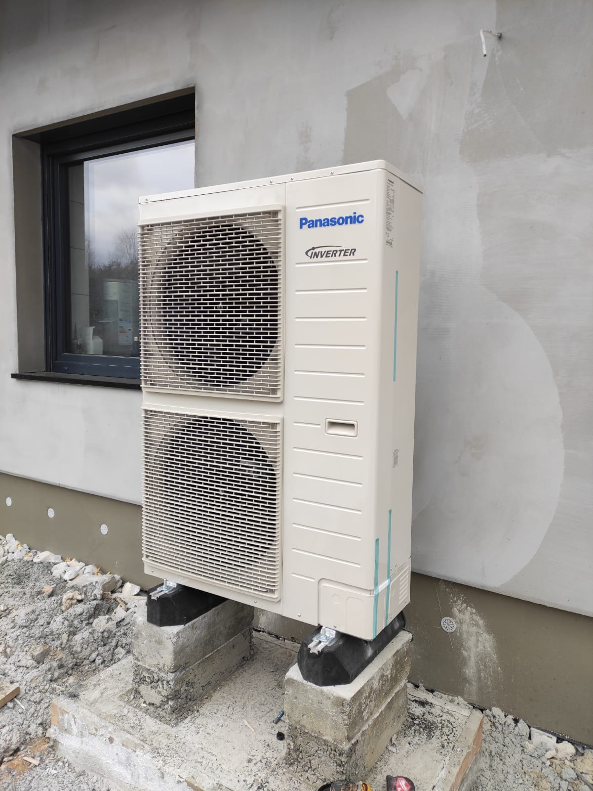Zewnętrzna jednostka pompy ciepła Panasonic Inverter zamontowana na betonowych blokach przy szarej ścianie budynku z widocznym oknem.
