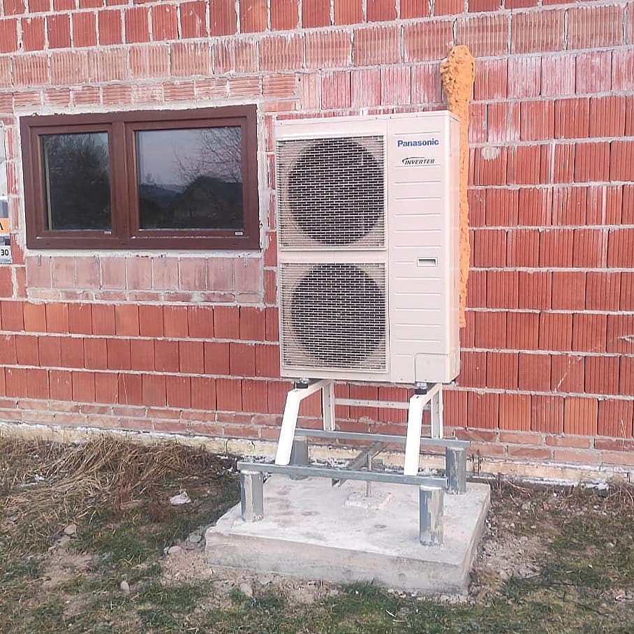 Zewnętrzna jednostka klimatyzacji Panasonic Inverter zamontowana na metalowej konstrukcji przy ścianie z czerwonej cegły, widoczne ślady pianki montażowej na ścianie i niedokończona elewacja.