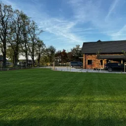 Rozległy, zadbany trawnik przed domem z cegły, z widocznymi drzewami i samochodami. Świeżo posadzona zieleń, równo skoszona trawa i błękitne niebo.