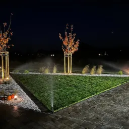 Ogród nocą z podświetlonymi drzewami w podporach, trawnikiem i systemem nawadniania. Ścieżka z kostki brukowej. Dekoracyjne oświetlenie i biały żwir.