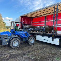 RozGardens Zakładanie i pielęgnacja ogrodów Patryk Różański - Operator w czerwonej czapce na niebieskim traktorze załadowuje paletę z rolkami trawy do czerwonej naczepy ciężarówki na tle zieleni i błękitnego nieba.