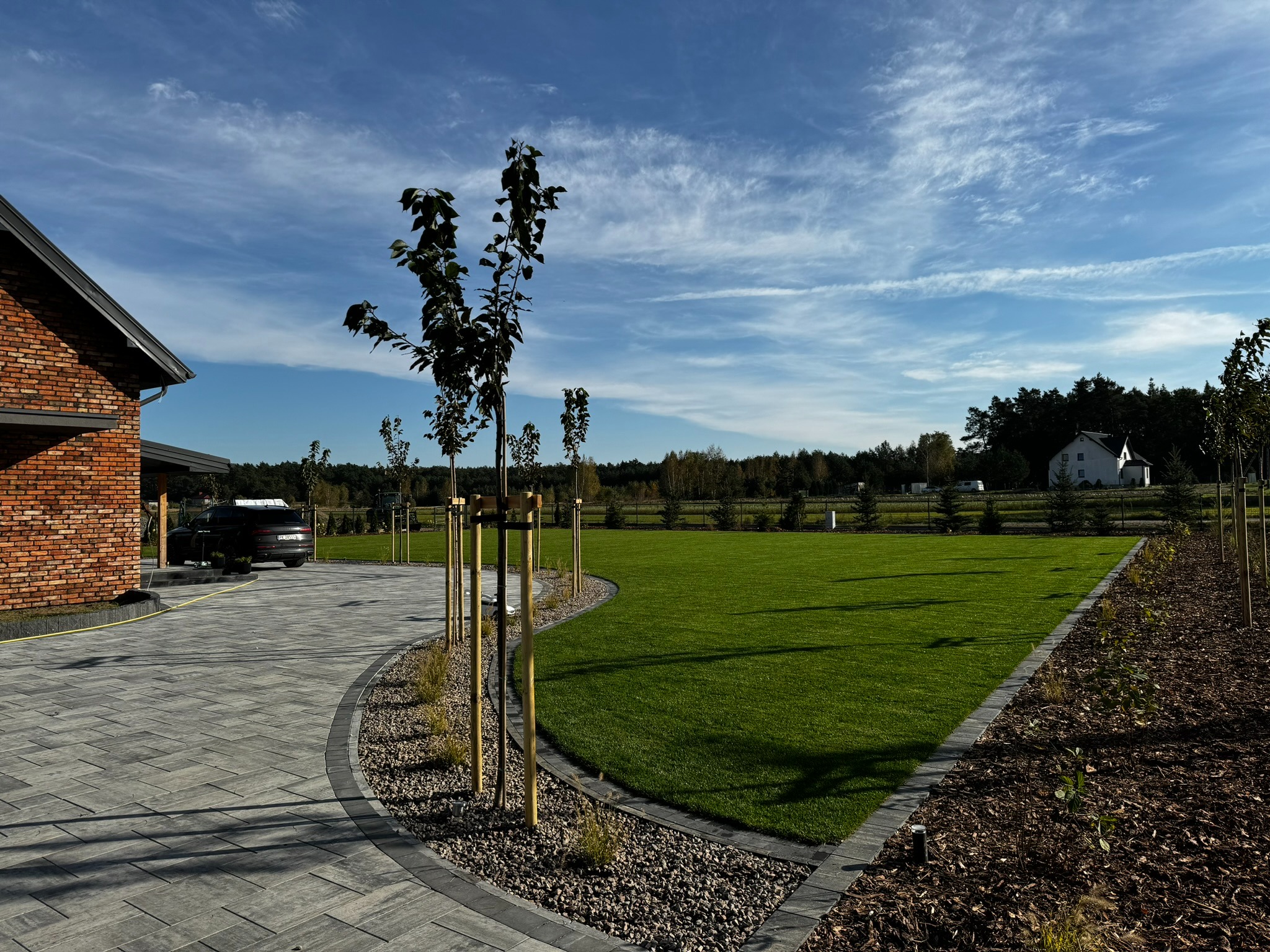 Nowoczesny ogród z zielonym trawnikiem, świeżo posadzone drzewa z podporami, kostka brukowa na podjeździe, dom z czerwonej cegły w tle, błękitne niebo.