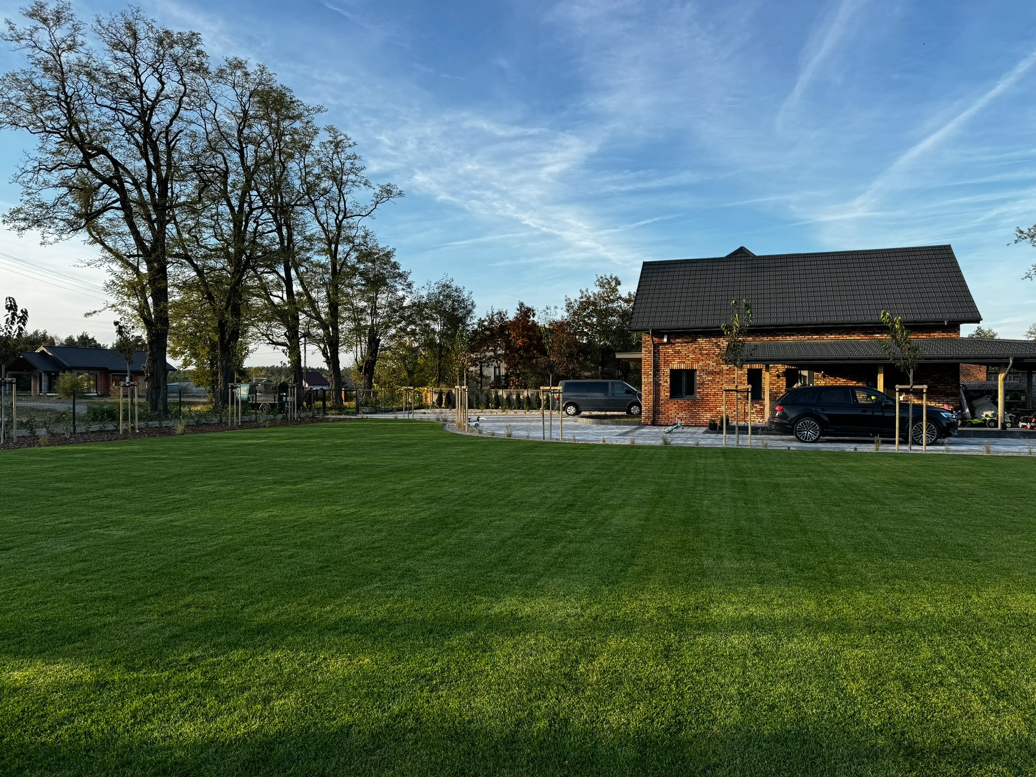 Rozległy, zadbany trawnik przed domem z cegły, z widocznymi drzewami i samochodami. Świeżo posadzona zieleń, równo skoszona trawa i błękitne niebo.