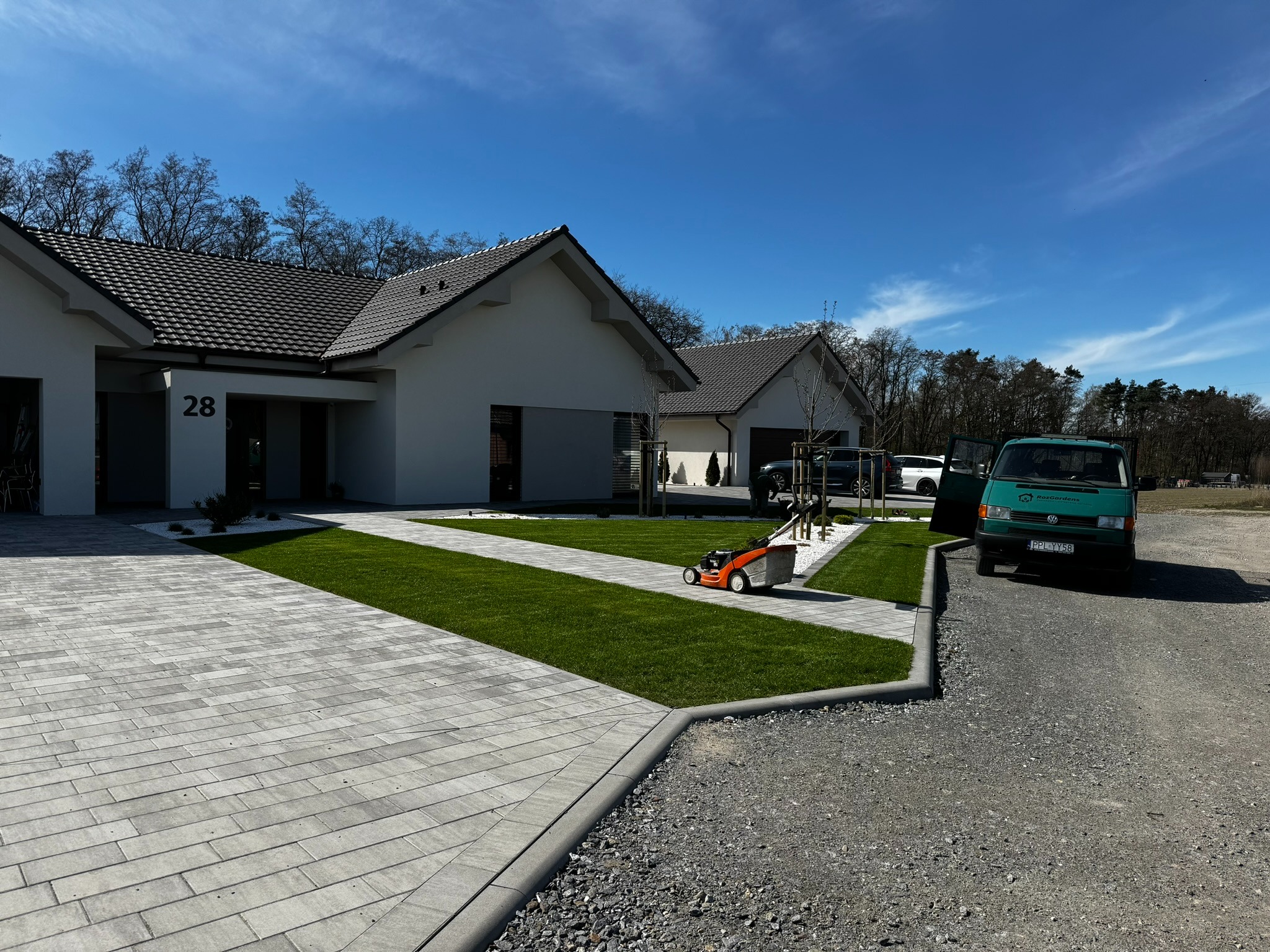 Nowoczesny dom z równo przystrzyżonym trawnikiem, kostką brukową i pomarańczową kosiarką. Zielony bus firmy ogrodniczej zaparkowany obok.