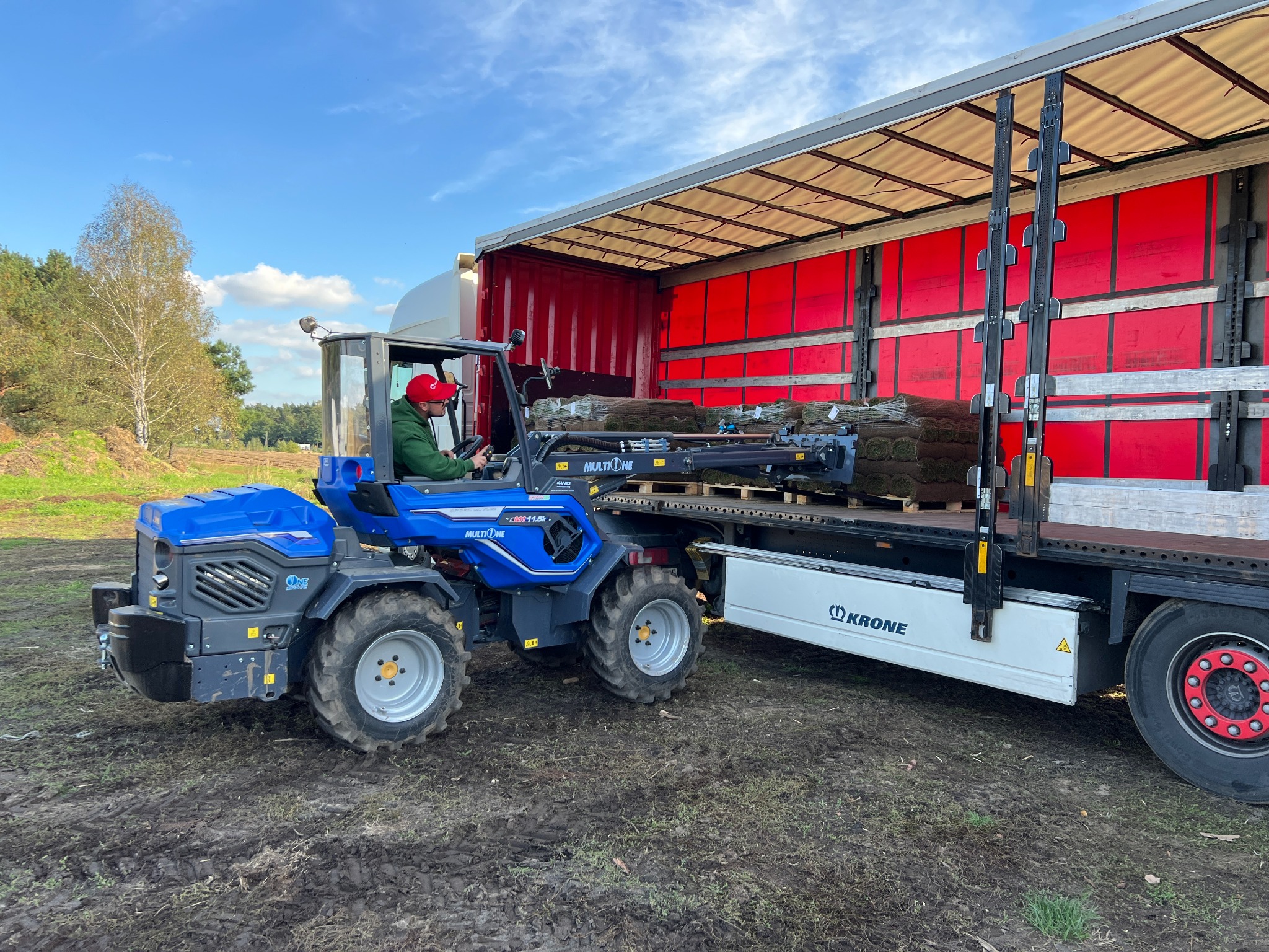 Operator w czerwonej czapce na niebieskim traktorze załadowuje paletę z rolkami trawy do czerwonej naczepy ciężarówki na tle zieleni i błękitnego nieba.