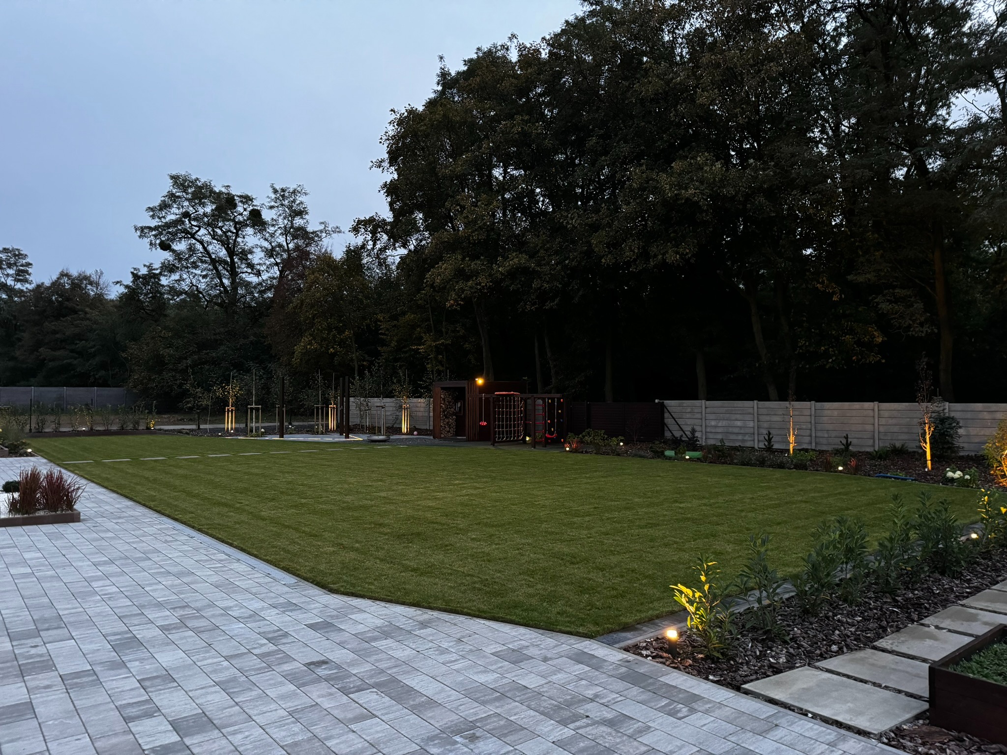 Zadbany, rozległy trawnik z równo przystrzyżoną trawą, obrzeża z kostki brukowej, oświetlenie punktowe, drzewa w tle. Widok na ogród w porze zmierzchu.