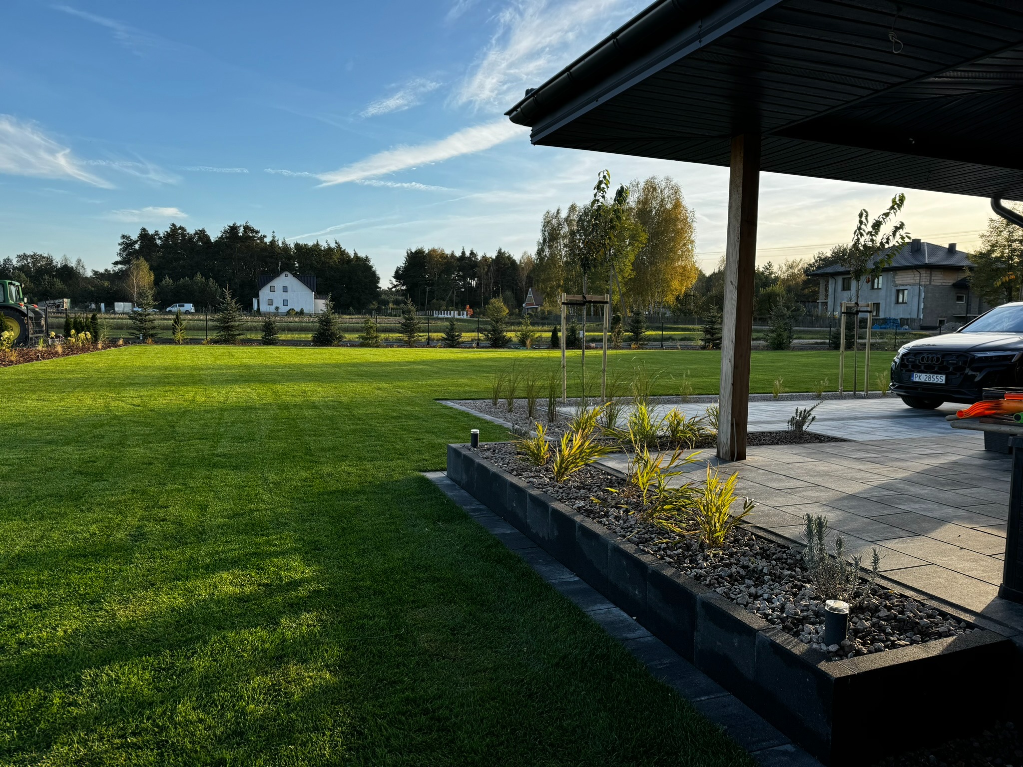 Zadbany ogród z równo przystrzyżonym trawnikiem, rabatami z kamieniami i roślinami ozdobnymi. W tle dom z białymi ścianami i drzewa. Widoczny fragment podjazdu z samochodem.