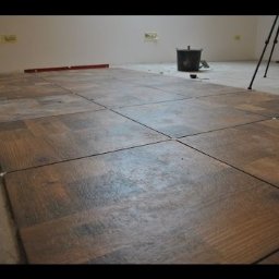 Przemysław Czerko - Układanie brązowych płytek podłogowych imitujących drewno, z białymi krzyżykami dystansowymi, poziomica na ścianie, wiadro z zaprawą w tle.