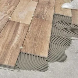 Układanie drewnopodobnych płytek podłogowych z użyciem krzyżyków dystansowych i świeżo nałożonej zaprawy klejowej, widoczne charakterystyczne żłobienia zaprawy.