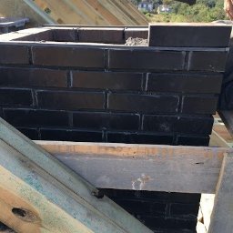 Przemysław Czerko - Budowa komina z czarnej cegły na dachu w Białej Podlaskiej, widoczne drewniane elementy konstrukcji dachowej i ręka murarza z kielnią.