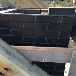 Budowa komina z czarnej cegły na dachu w Białej Podlaskiej, widoczne drewniane elementy konstrukcji dachowej i ręka murarza z kielnią.