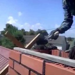 Przemysław Czerko - Murarz w koszuli w kratę, układający cegły na dachu, używając kielni i poziomicy, na tle błękitnego nieba i zieleni.