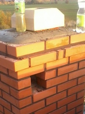 Ceglany komin w trakcie budowy, z widocznym otworem wentylacyjnym i butelkami z napojami na szczycie.