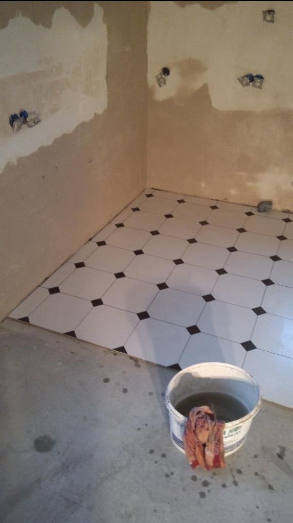 Układanie białych płytek podłogowych z czarnymi akcentami w rogu pomieszczenia, widoczny kubeł z brudną wodą i szmatą, ściany przygotowane do dalszych prac.