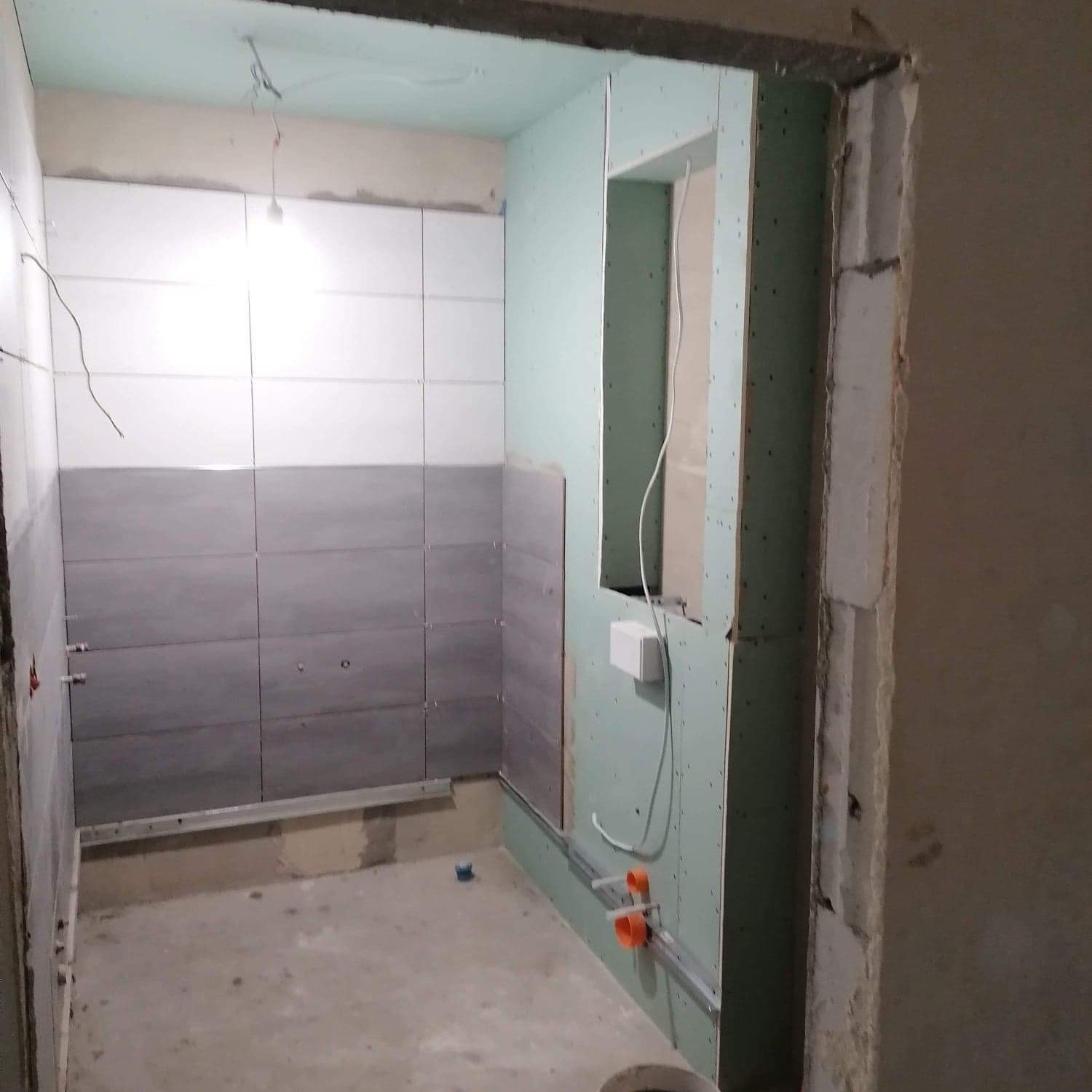 Wnętrze łazienki w trakcie remontu: częściowo ułożone płytki ceramiczne w odcieniach szarości i bieli, widoczne profile pod płyty gipsowo-kartonowe, wystające przewody elektryczne i instalacja wodna.