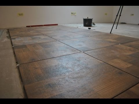 Układanie brązowych płytek podłogowych imitujących drewno, z białymi krzyżykami dystansowymi, poziomica na ścianie, wiadro z zaprawą w tle.