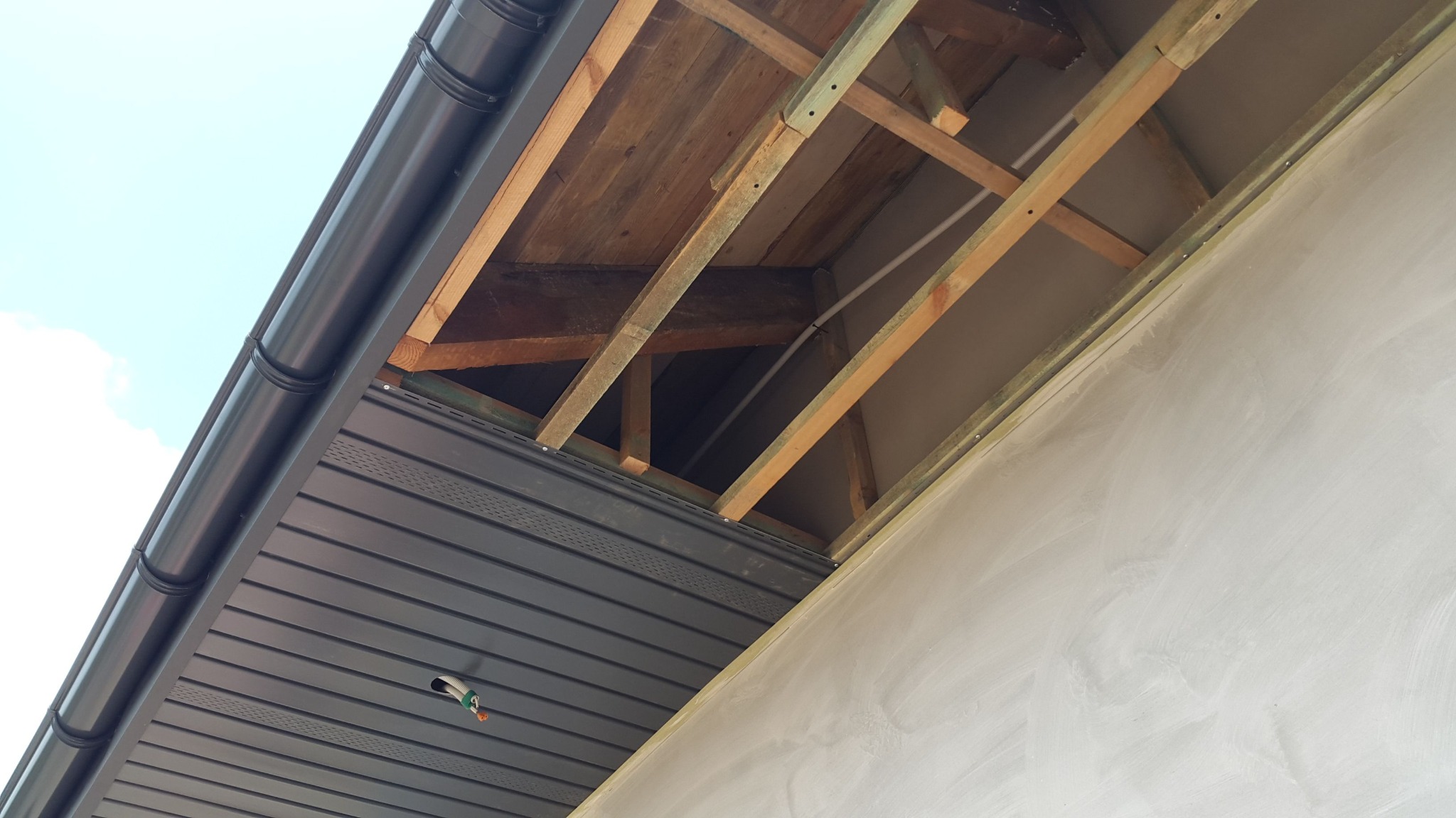Podbitka dachowa z szarego panelu PCV z widocznym systemem rynnowym, drewnianą konstrukcją dachu i fragmentem otynkowanej ściany budynku w trakcie montażu.
