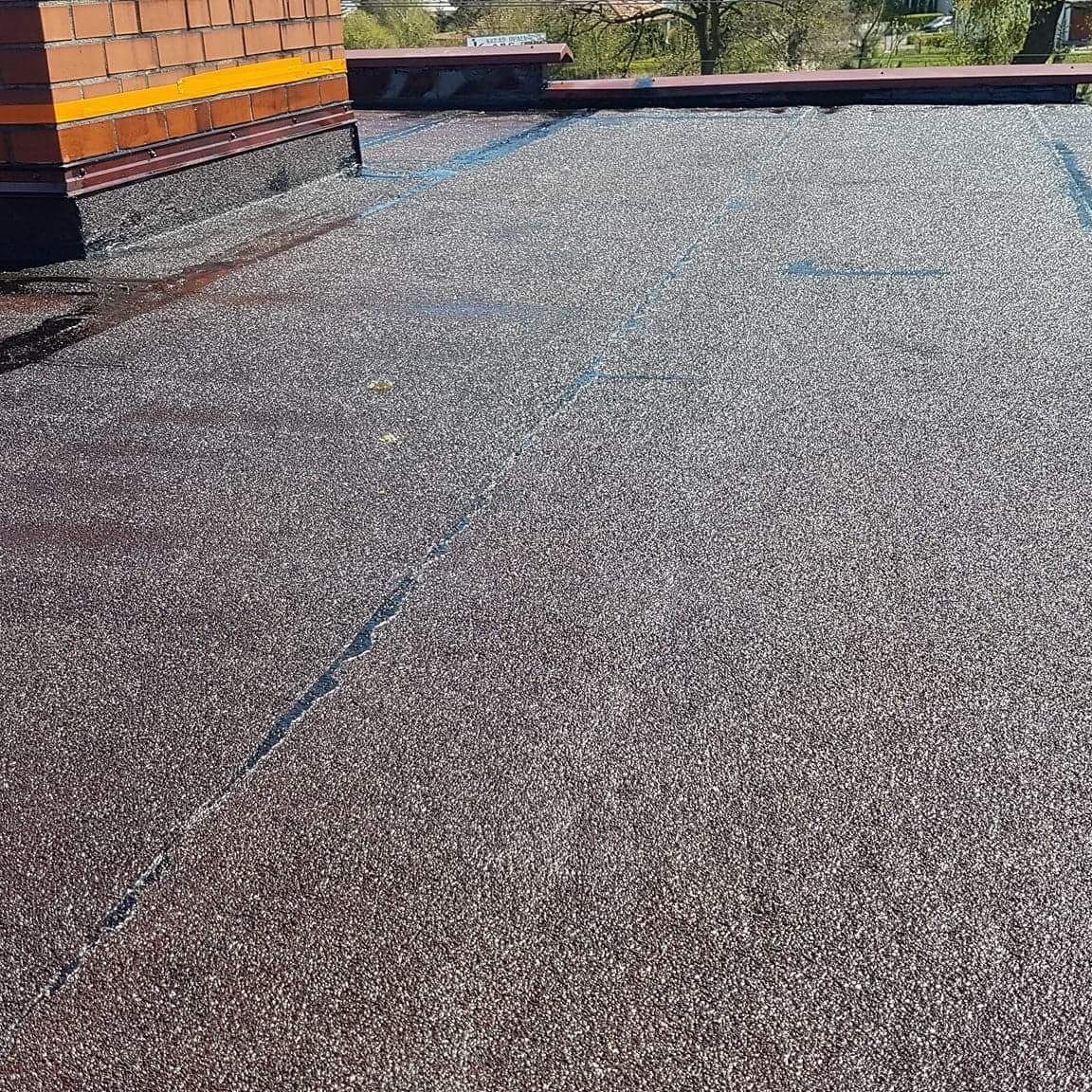 Widok z góry na płaski dach pokryty papą z widocznym kominem i śladami wilgoci wzdłuż łączeń.