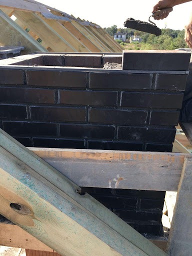 Budowa komina z czarnej cegły na dachu w Białej Podlaskiej, widoczne drewniane elementy konstrukcji dachowej i ręka murarza z kielnią.