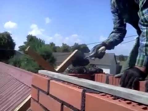 Murarz w koszuli w kratę, układający cegły na dachu, używając kielni i poziomicy, na tle błękitnego nieba i zieleni.