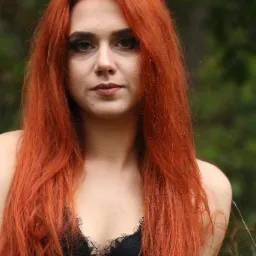 Portret kobiety z długimi, rudymi włosami i czarną koronkową bielizną na tle zieleni.