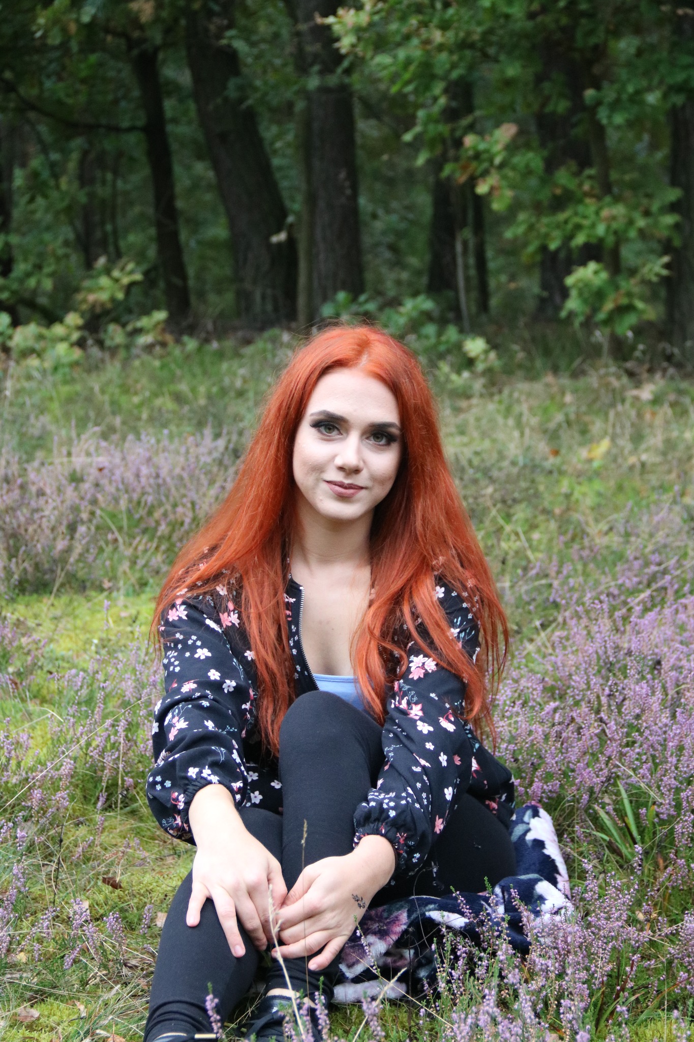 Portret kobiety z rudymi włosami siedzącej na wrzosowisku w lesie, ubrana w czarną bluzkę w kwiaty i czarne legginsy, z delikatnym uśmiechem.