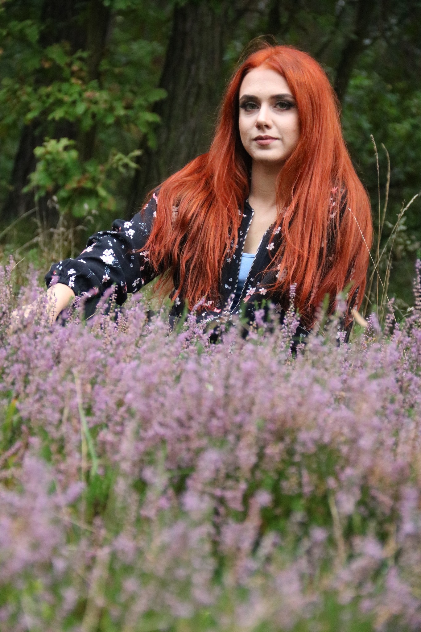Kobieta z długimi rudymi włosami w kwiecistej kurtce stoi w polu fioletowych wrzosów, z zamyślonym wyrazem twarzy i lasem w tle.