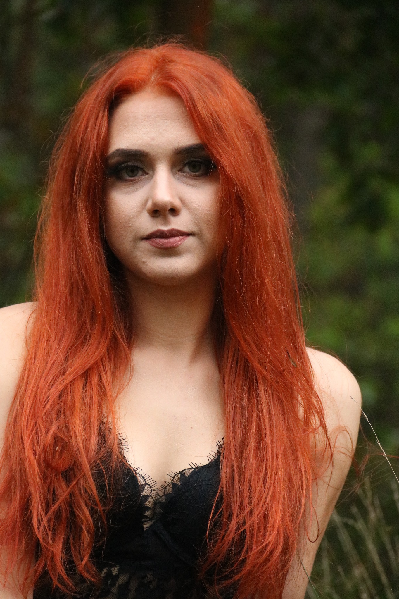Portret kobiety z długimi, rudymi włosami i czarną koronkową bielizną na tle zieleni.