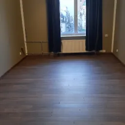 Pusta, odnowiona przestrzeń z ciemnymi panelami podłogowymi, brązowymi ścianami i oknem z widokiem na miasto, zasłoniętym ciemnymi zasłonami.