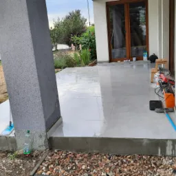 Świeżo wyłożony, mokry taras z szarych płytek, widoczny słupek podtrzymujący z niedoczyszczonymi resztkami zaprawy, narzędzia i akcesoria do czyszczenia w rogu tarasu.