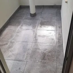 Balkon po położeniu szarych, kwadratowych płytek podłogowych; widoczna kolumna nośna i butelka środka czyszczącego na parapecie.