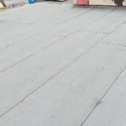 Szary, płaski dach pokryty papą termozgrzewalną z widocznymi łączeniami, kominem i elementami instalacji na tle nieba.