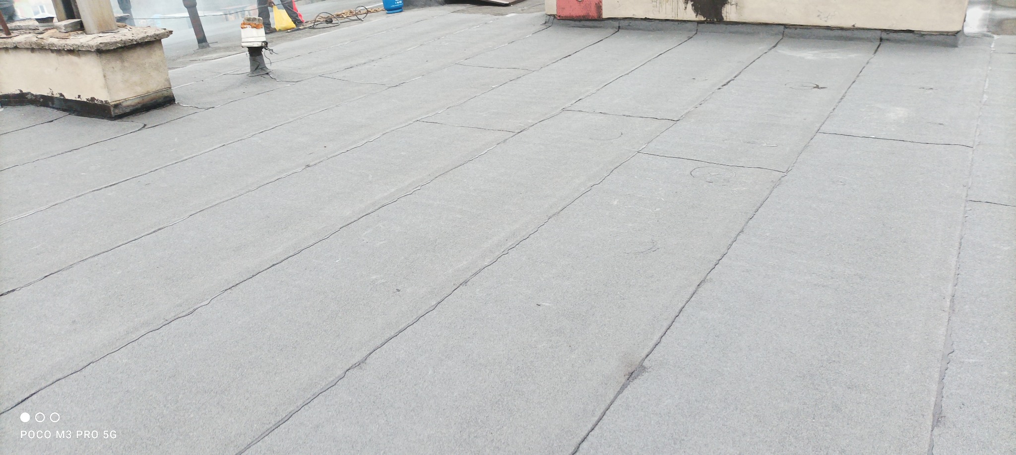 Szary, płaski dach pokryty papą termozgrzewalną z widocznymi łączeniami, kominem i elementami instalacji na tle nieba.