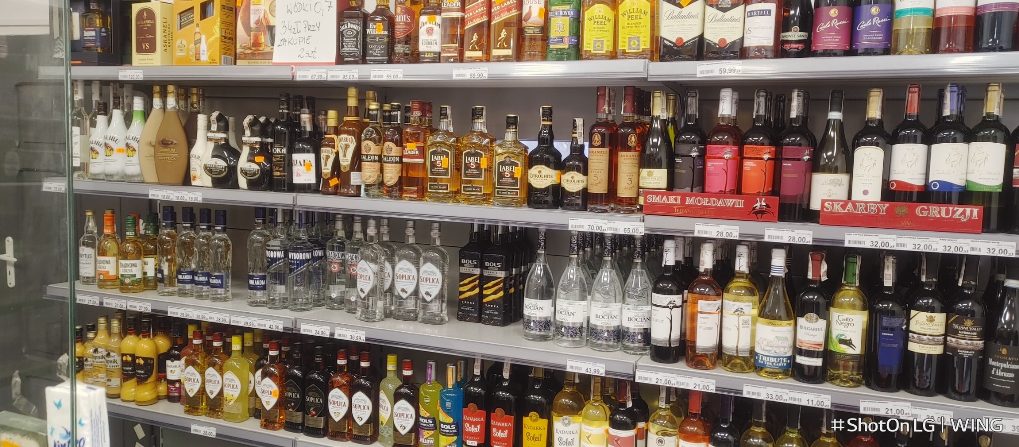 Półki sklepowe wypełnione różnorodnymi butelkami alkoholi, od wódek i likierów po whisky i wina, z widocznymi cenami i etykietami w zbliżeniu.