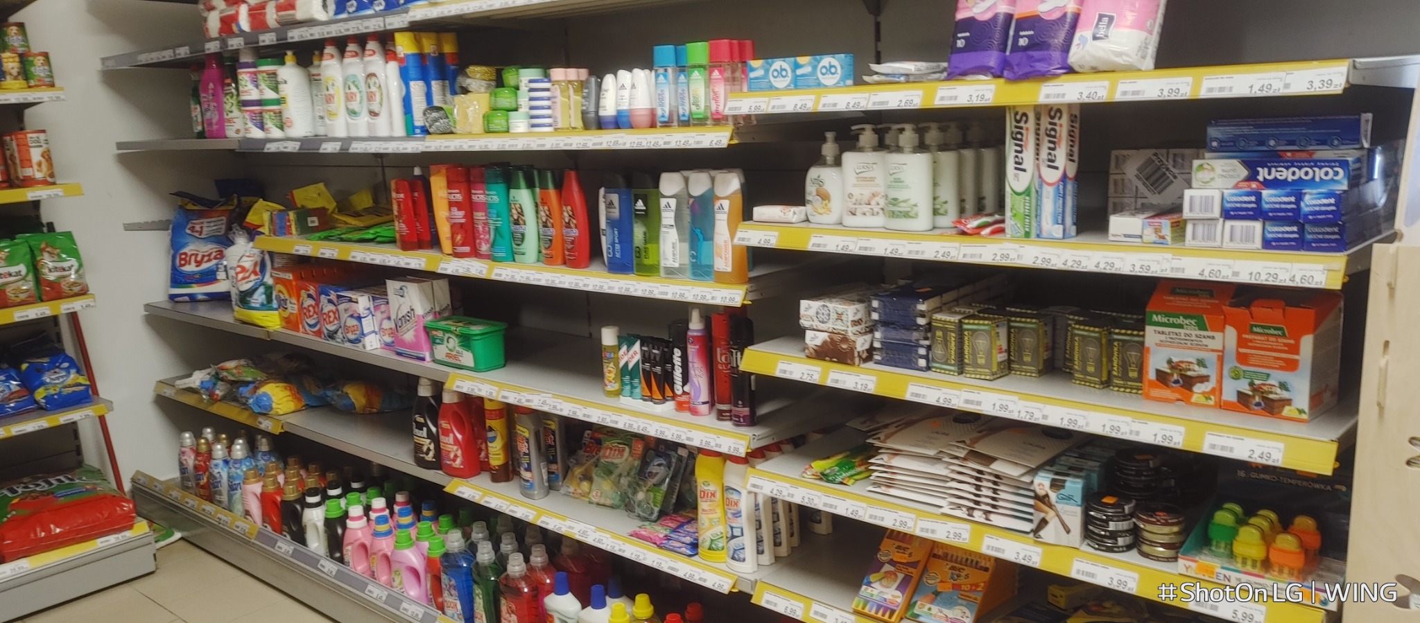 Półki w sklepie z szerokim wyborem środków czystości, kosmetyków i artykułów higienicznych, w tym proszków do prania, płynów do mycia naczyń, szamponów, mydeł i past do zębów, z widocznymi cenami...