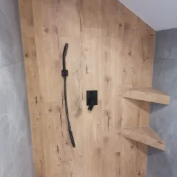 Nowoczesna kabina prysznicowa z drewnopodobną okładziną, czarną armaturą i narożnymi półkami z tego samego materiału.