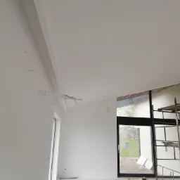 Wnętrze w trakcie malowania: świeżo pomalowane ściany na biało, widoczne przewody elektryczne wystające ze ściany, okno z czarną ramą i rusztowanie.