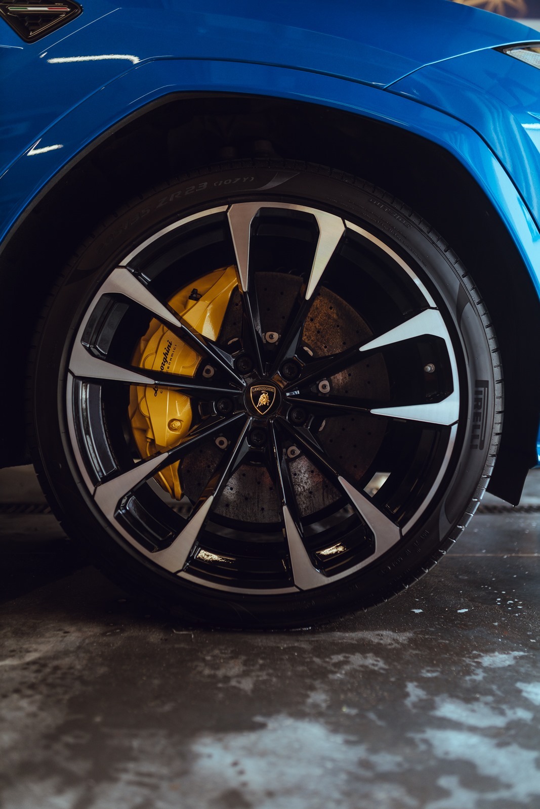 Błyszczące, niebieskie nadwozie Lamborghini Urus z widocznym kołem z czarną felgą o srebrnych akcentach i żółtym zaciskiem hamulcowym Brembo z logo Lamborghini, stojące na mokrej posadzce.