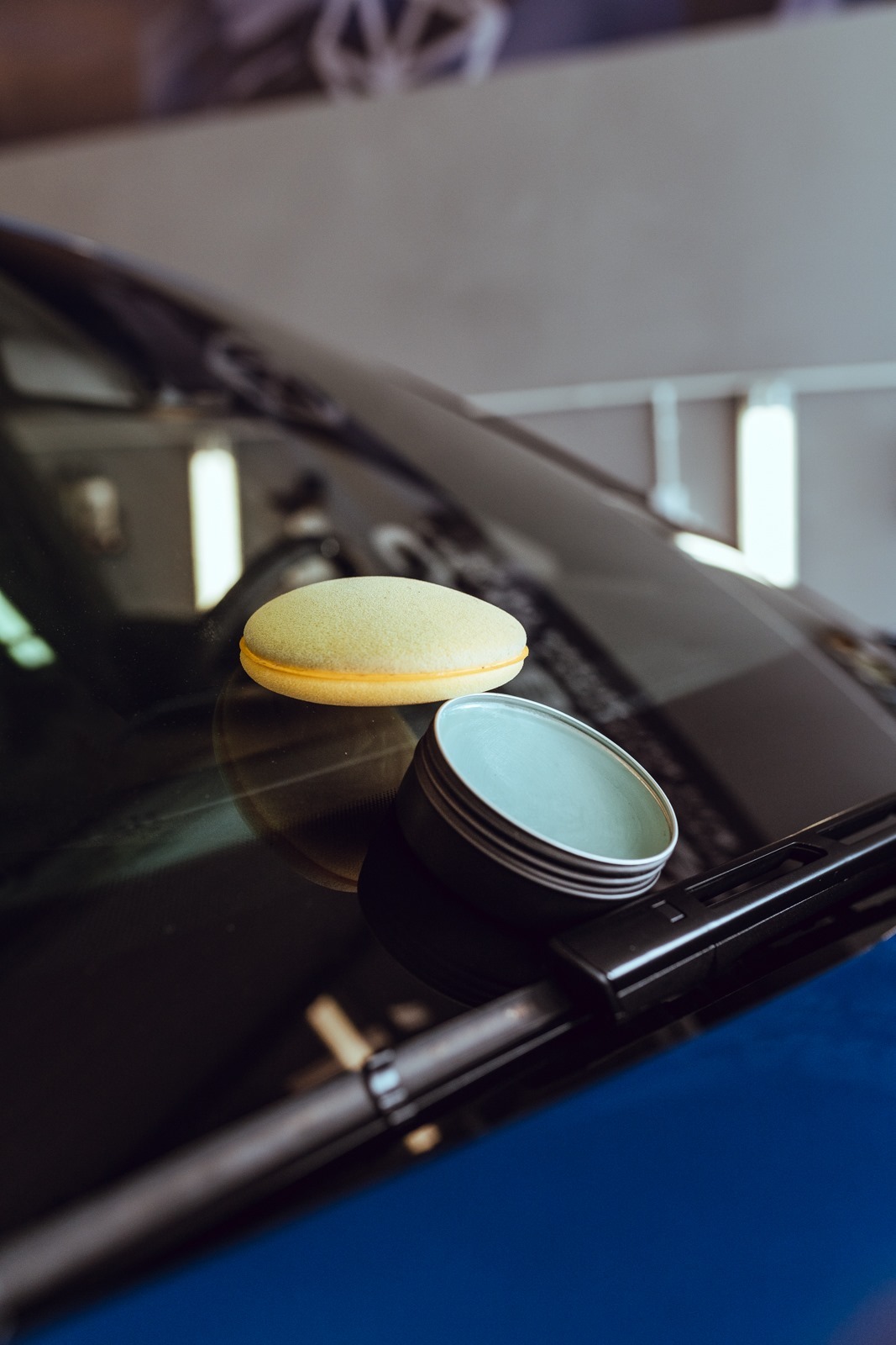 Żółta gąbka polerska i puszka z woskiem na przedniej szybie niebieskiego samochodu, widoczne wycieraczki.
